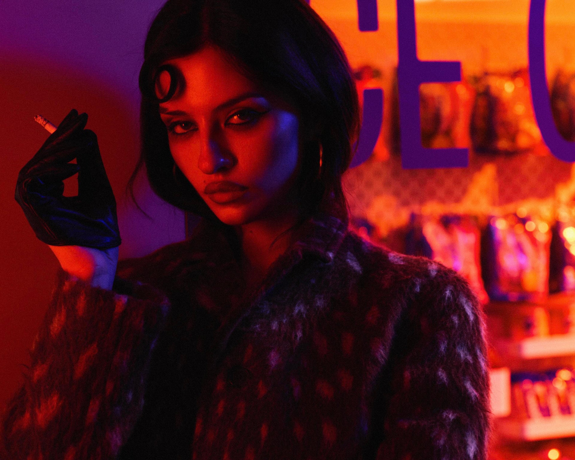 Person with dark hair and black gloves holding a cigarette in a neon-lit setting